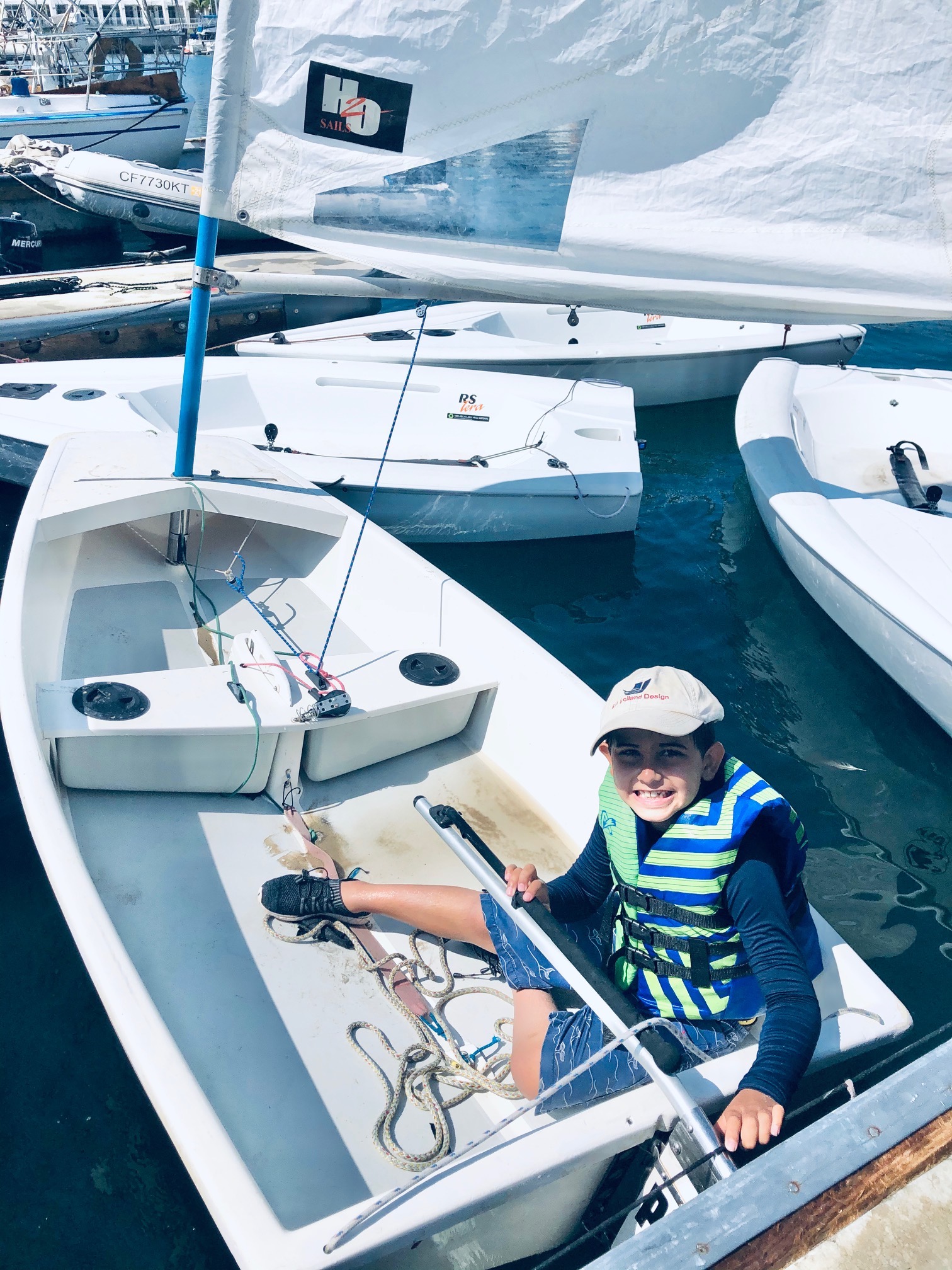 Wearing a Ron Holland Design hat young sailor ready to sail as he sits in his boat among a flotilla